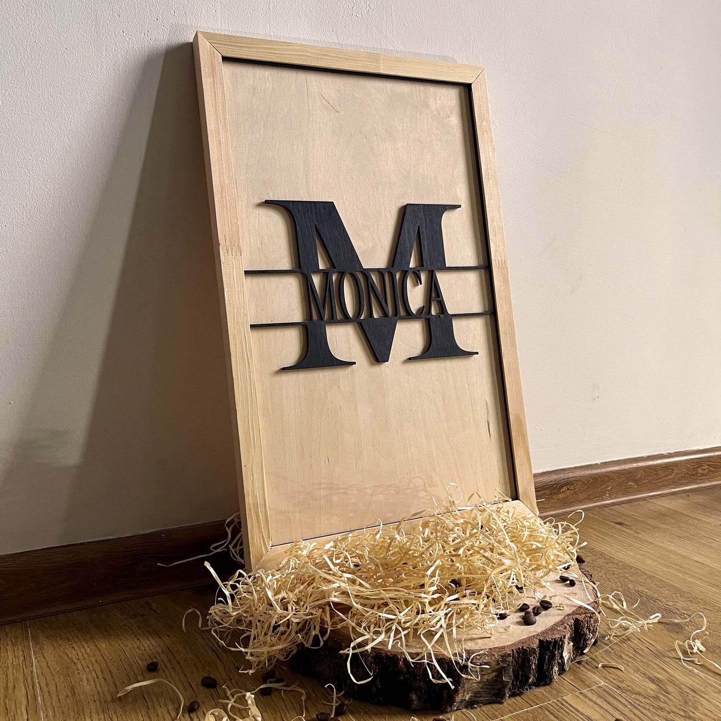 MONOGRAM - Ręcznie wykonany drewniany obraz przedstawiający czarną dekoracyjną literę M na jasnym drewnianym tle, oprawiony w naturalne drewno, wyeksponowany na rustykalnym kawałku drewna ze słomianą dekoracją. Elegancki spersonalizowany element wystroju domu.